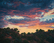 Painting by Brenda Howell of dramatic colorful clouds as sun sets above juniper and pinon covered hills in Northern New Mexico.